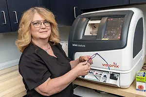 A woman in a black shirt with blonde hair stands in front of a Visionix machine, holding glasses.