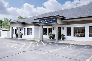 A vintage optical building with a grey roof, white walls, and a blue sign that says Vintage Optical. There are potted plants in front of the entrance, and yellow markings are on the parking lot.