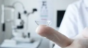 A close up of a finger holding a contact lens in front of a blurry eye chart.