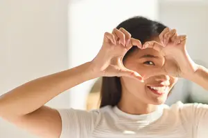 A woman smiling and making a heart shape with her hands in a room