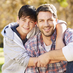 A father and son pose in a park with trees in the background.
