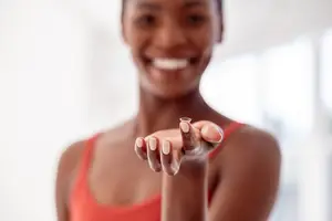 A woman wearing a red sleeveless top holds a small contact lens in her right hand and smiles at the camera.