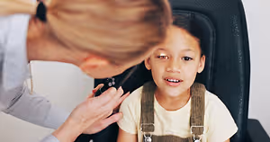 A woman, likely a doctor, is examining a young girl's ear with a medical instrument, both are wearing casual clothing.