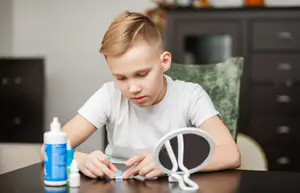 A young boy is sitting at a table, holding a bottle of glue and a small white bottle, and applying glue to a dental brace. He is looking at a mirror on the table. Behind him, there is a cabinet with drawers and a pumpkin on top. There is a chair on the right side of the image.