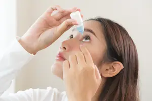A woman in a white long-sleeve shirt is applying eye drops to her eye.
