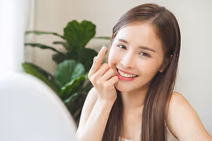 A woman with long hair is sitting and smiling while adjusting her contact lens in front of a mirror. She is wearing a sleeveless shirt and is holding her cheek with her left hand. Behind her, there is a plant with green leaves, and the wall is painted white. The woman's reflection is also visible in the mirror.