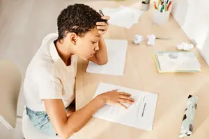 A child is sitting on a chair, leaning on a wooden desk, reading a piece of paper with a pen in their hand. The desk has papers, a pen holder, and a potted plant. Behind the child is a white wall. The child is wearing a white shirt and blue shorts.