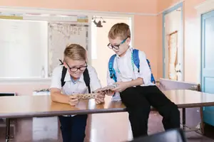 Two young boys in school uniforms and glasses, examining something on a desk in a classroom.