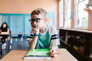 A young boy sitting at a desk in a classroom, wearing glasses, and looking at a book on the desk in front of him.
