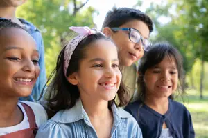 Five smiling children standing in a park with trees in the background