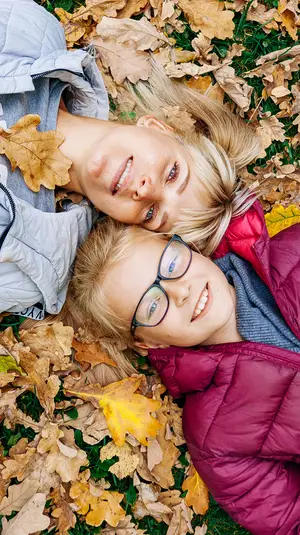 A woman and a young girl are lying on the grass, surrounded by autumn leaves, both wearing warm clothing and smiling.