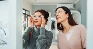 Two women wearing glasses are standing in a room and looking at something. One of them is holding her face with both hands, and the other is holding her glasses with her left hand. Behind them is a glass door with a reflection of a person.