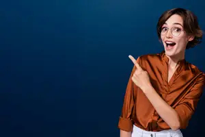 A woman wearing a brown blouse and glasses is smiling and pointing at something while standing in front of a blue background.
