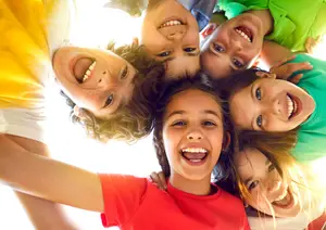A group of children are standing in a circle and smiling with their mouths open and their arms around each other.