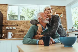 A couple is sitting on a chair in a kitchen, smiling and looking at each other while embracing each other. The kitchen is well-lit and has a brick wall, a wooden cabinet, a wooden desk, a teapot, and a kettle. Outside, there is a bush and a tree. The woman is wearing glasses and a sweater, while the man is wearing a polo shirt and a necklace.