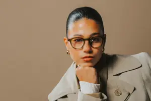 A woman with glasses and a ring on her finger wearing a trench coat and a white shirt