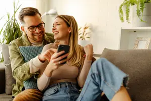 A man and a woman are sitting on a couch and holding a smartphone and smiling while looking at it