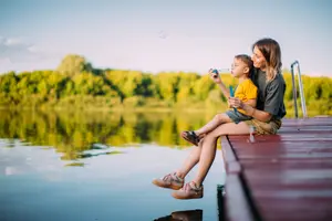 A woman and a child are sitting on a dock by a lake, and the child is blowing bubbles into the air