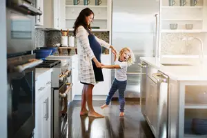 A pregnant woman and a child are standing in the kitchen and seem to be playing together. They are standing on the wooden floor. A refrigerator, a cabinet, and a sink are on the right side, and a gas stove is on the left. A bowl, cups, plates, and a pot are placed on the shelf. A microwave oven is in the corner. The reflection of the gas stove is reflected on the floor.