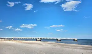 A white bridge over a calm body of water with a sandy shore and a blue sky with clouds in the background.