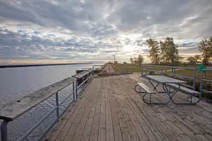 A wooden deck with benches and a picnic table overlooking a lake and a grassy area with trees under a cloudy sky.