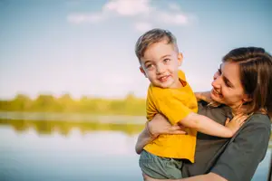 A smiling young boy in a yellow t-shirt is being held by his mother, standing near a calm lake with trees in the background under a blue sky with some clouds.