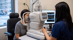 An older woman with short hair wearing a mask is getting her eyes examined by a woman in a blue shirt.