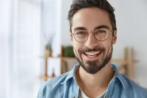 A man wearing glasses and a blue shirt is smiling while standing in front of a wooden shelf with plants and a white curtain.