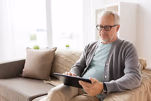 An older man sits on a couch using a tablet in a living room