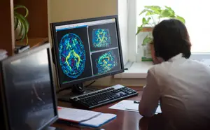 A person sitting at a desk in front of a computer screen with a brain image displayed