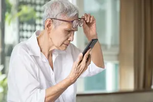 An elderly woman in a white shirt adjusts her glasses while looking at her phone.