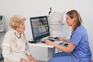 An elderly woman and a woman in blue scrubs sitting in front of a computer monitor with a medical device behind them.