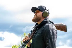A man wearing a hat, glasses, and headphones is holding a shotgun and looking up at the sky. He is wearing a jacket, pants, and a watch. He is probably hunting in a field with green trees and plants. The sky is blue and has some clouds.