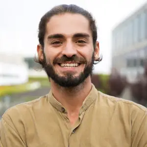 Smiling man with a beard and mustache wearing a brown shirt standing outside near a building