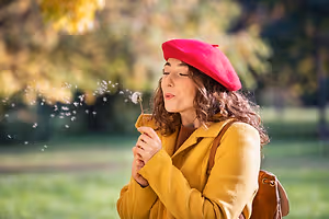 A woman wearing a red beret and yellow coat blowing on a dandelion