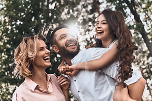 A man is holding a young girl, and a woman is smiling and looking at them outside in the park