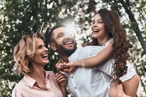 A man is holding a young girl, and a woman is smiling and looking at them outside in the park