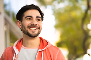 A smiling man wearing a red jacket with white linings on the collar and sleeves. He is outdoors in a well-lit area with a building and trees in the background.