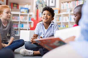 Four kids sitting on the floor in a library reading books and smiling