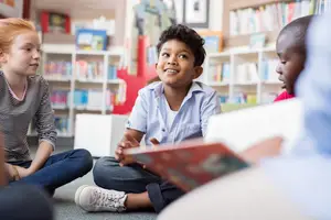 Four kids sitting on the floor in a library reading books and smiling