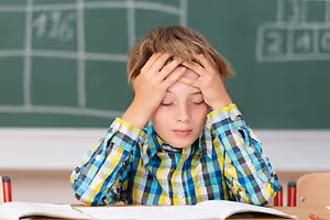 A young boy sitting in a classroom, holding his head with both hands and looking stressed.