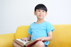 A smiling young boy with black hair sits on a yellow couch, holding a book in both hands.