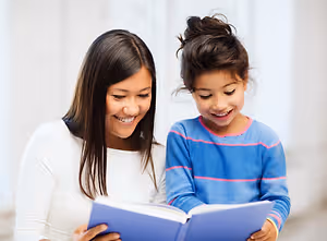A woman and a girl are sitting together and reading a book. The woman is holding the book, and the girl is looking at it.