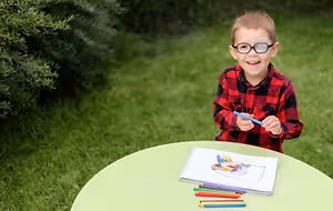 A little boy is smiling while sitting on a table and holding a blue pen in his hand. He is wearing a red and black checkered shirt and glasses. There is a green table with a notebook, a pen, and a pencil on it. There are also some colored pencils on the table. Behind him is a green lawn with some plants and bushes.