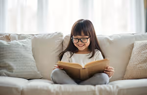 A young girl wearing glasses sits on a couch and reads a book while smiling.