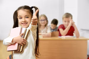 A girl holding books with a thumb up and a few other kids sitting behind her in a classroom.