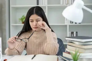 A woman wearing a beige turtleneck sweater sits at a desk with books, a lamp, and a potted plant, holding her glasses and looking thoughtful.