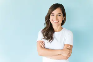 A woman with brown hair wearing a white t-shirt is smiling and posing for a photo with her arms crossed on a blue background.