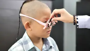 A young boy with a shaved head and a bracelet on his wrist is getting an eye exam by a doctor wearing a white coat and bracelet.