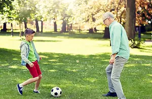 An older man wearing glasses and a young boy in a green shirt and red shorts are playing soccer in the park.